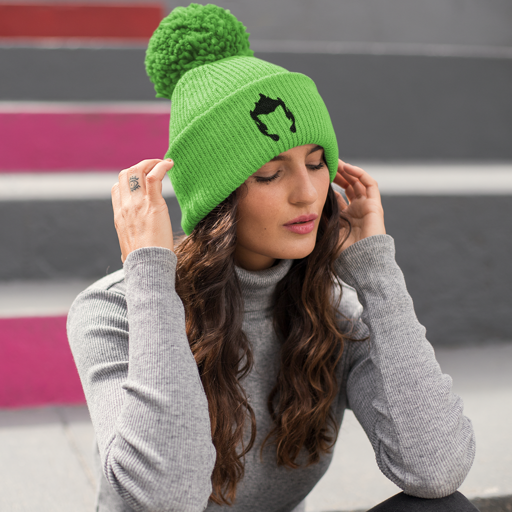 a woman sitting on the ground wearing a green beanie with a pom pom and the fire kitty embroidered design by diskittysonfire