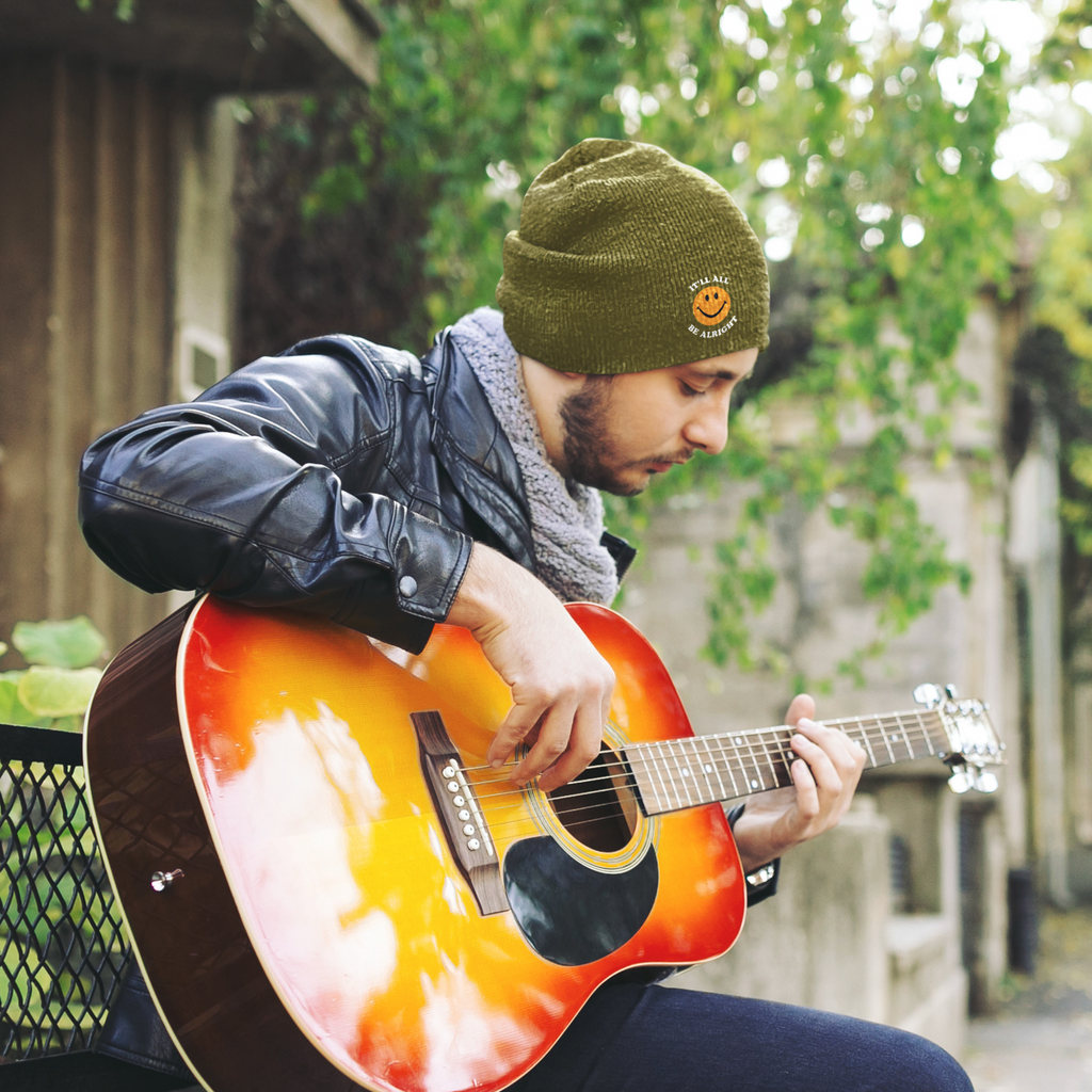 man playing guitar and wearing green beanie with smiley alright logo