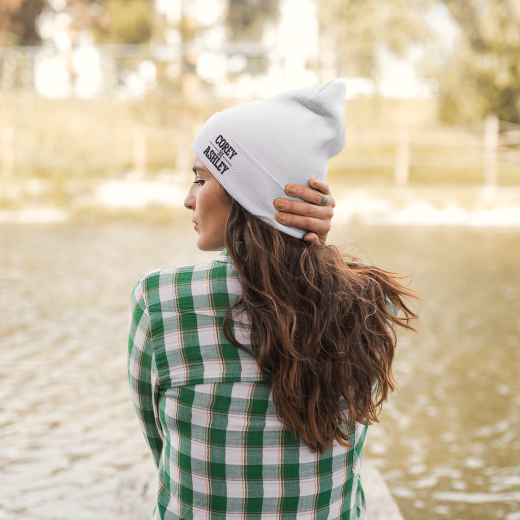 a woman sitting in front of a lake with a white beanie with the corey and ashley wordmark