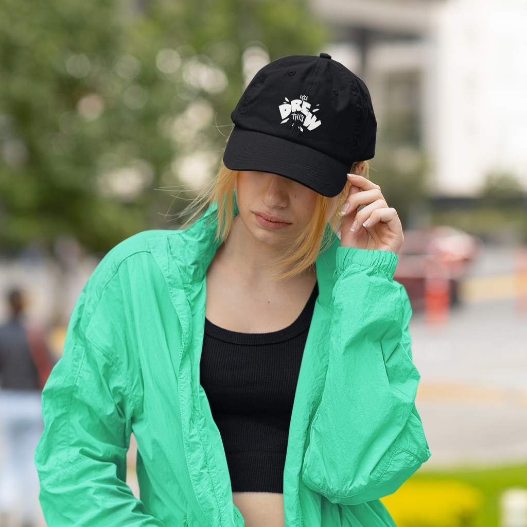 a woman wearing a green wind breaker and a black dad hat with the letsdrewthis logo embroidered on the front