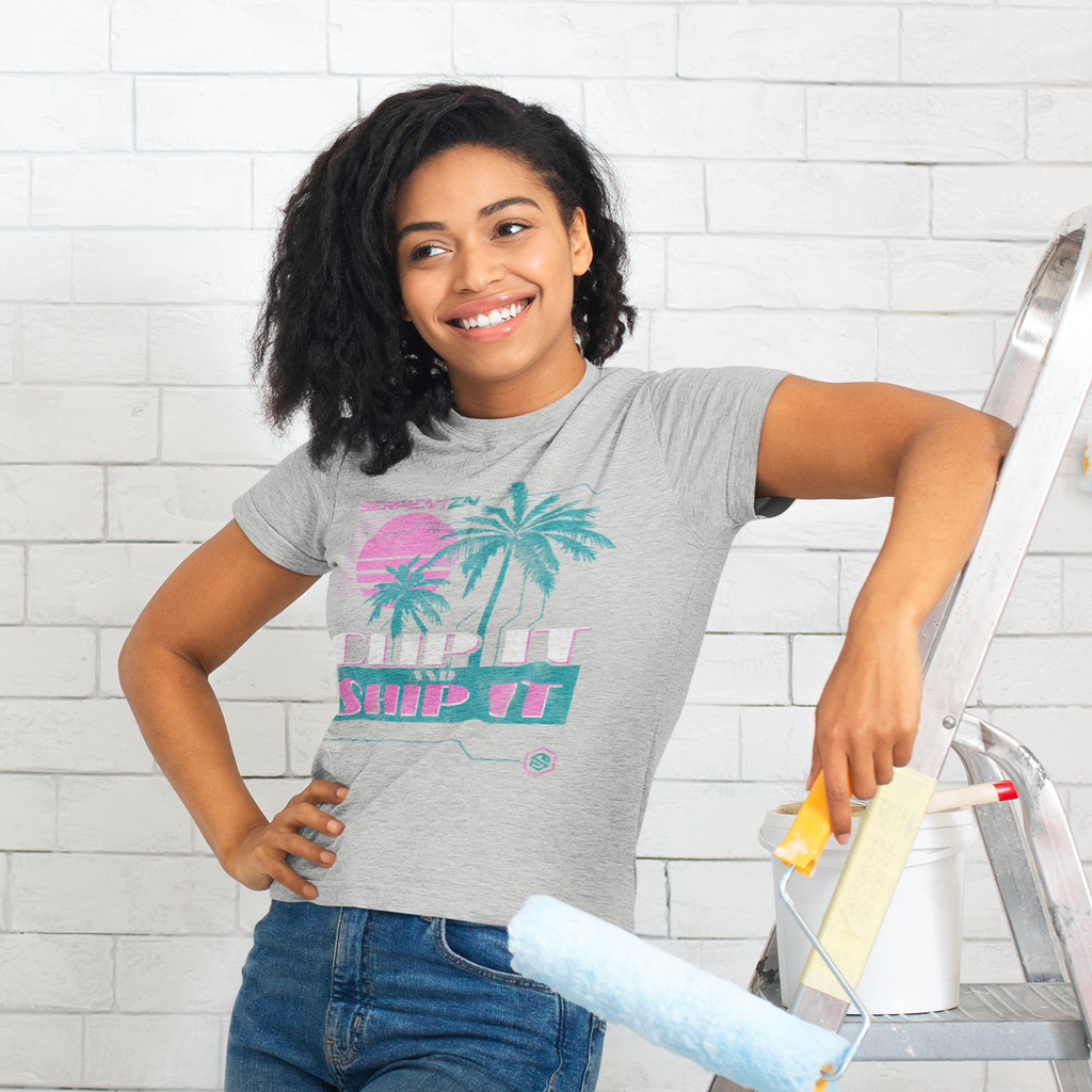 a woman leaning against a ladder and holding a paint roller.  She is wearing an athletic heather tshirt with the clip it and ship it design from serpentzn.