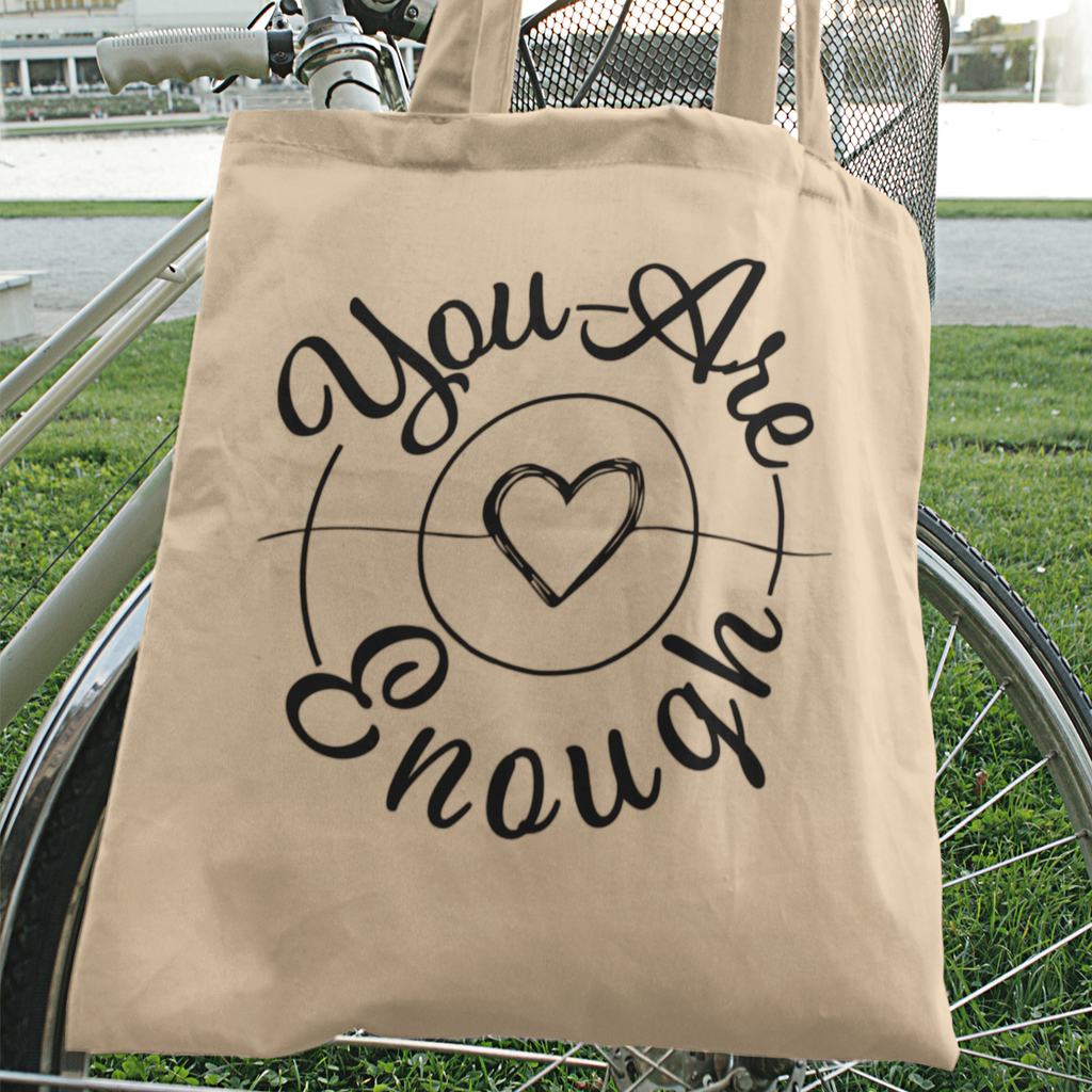 A tote bag with the mystiraynes you are enough design on it hanging from a bicycle 
