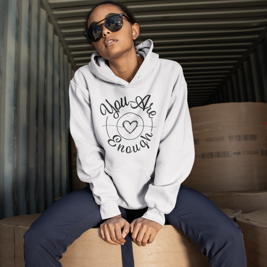 A woman wearing a white hoodie, sitting on boxes in a shipping container, wearing the mystiraynes You Are Enough Design