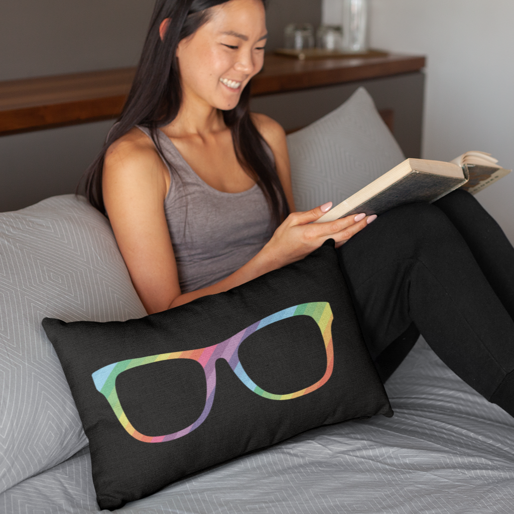 A woman reading a book on a bed with the megggg rainbow glasses pillow next to her.