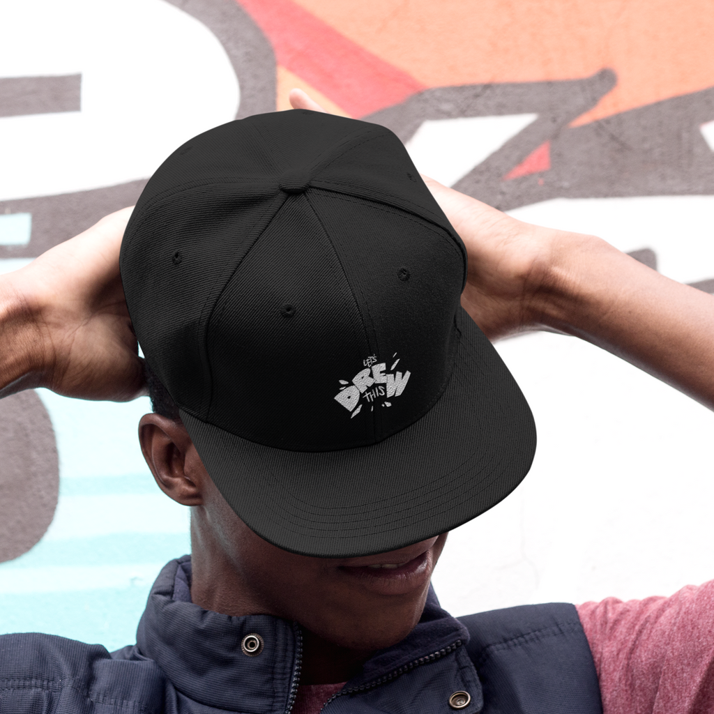 a man wearing a black snapback and holding it over his eyes.  the snapback has the LetDrewIt logo embroidered on it.