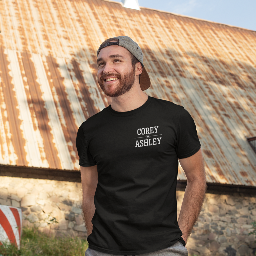 a man standing in front of a barn with a black hat and the corey and ashley wordmark on the left chest