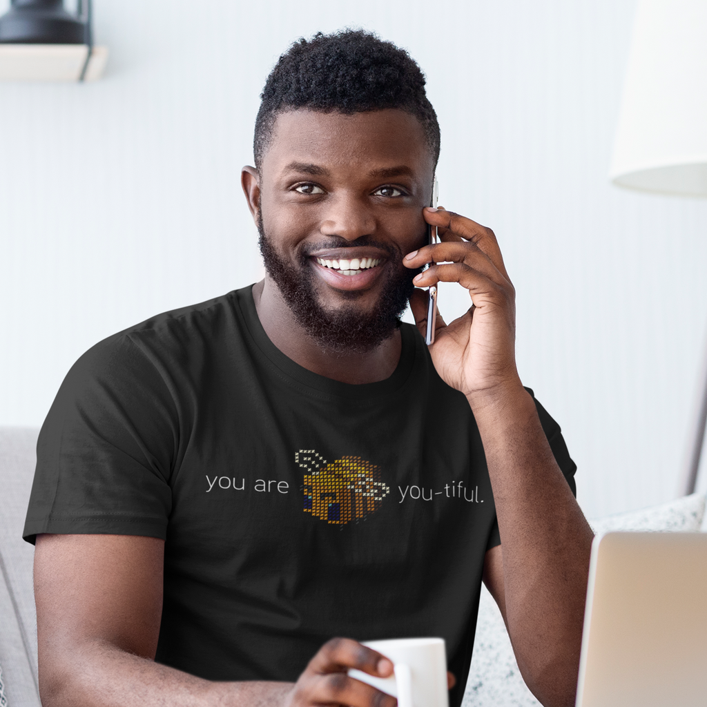 A man with a beard holding a cell phone to his ear and wearing a black shirt with the You Are Bee-You-Tiful design.