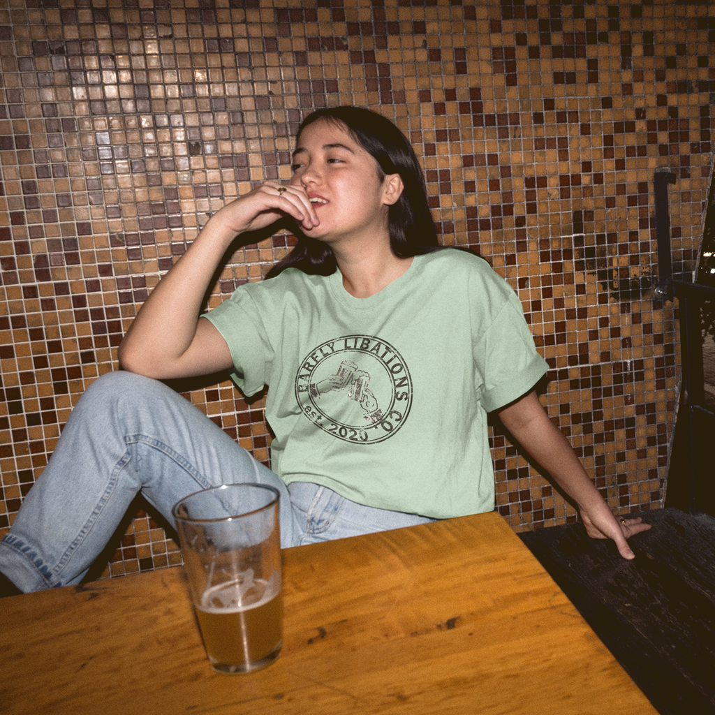 a woman sitting in a bar with a beer wearing a heather prism mint tshirt with the Barfly Libations Company logo from kisogoplays