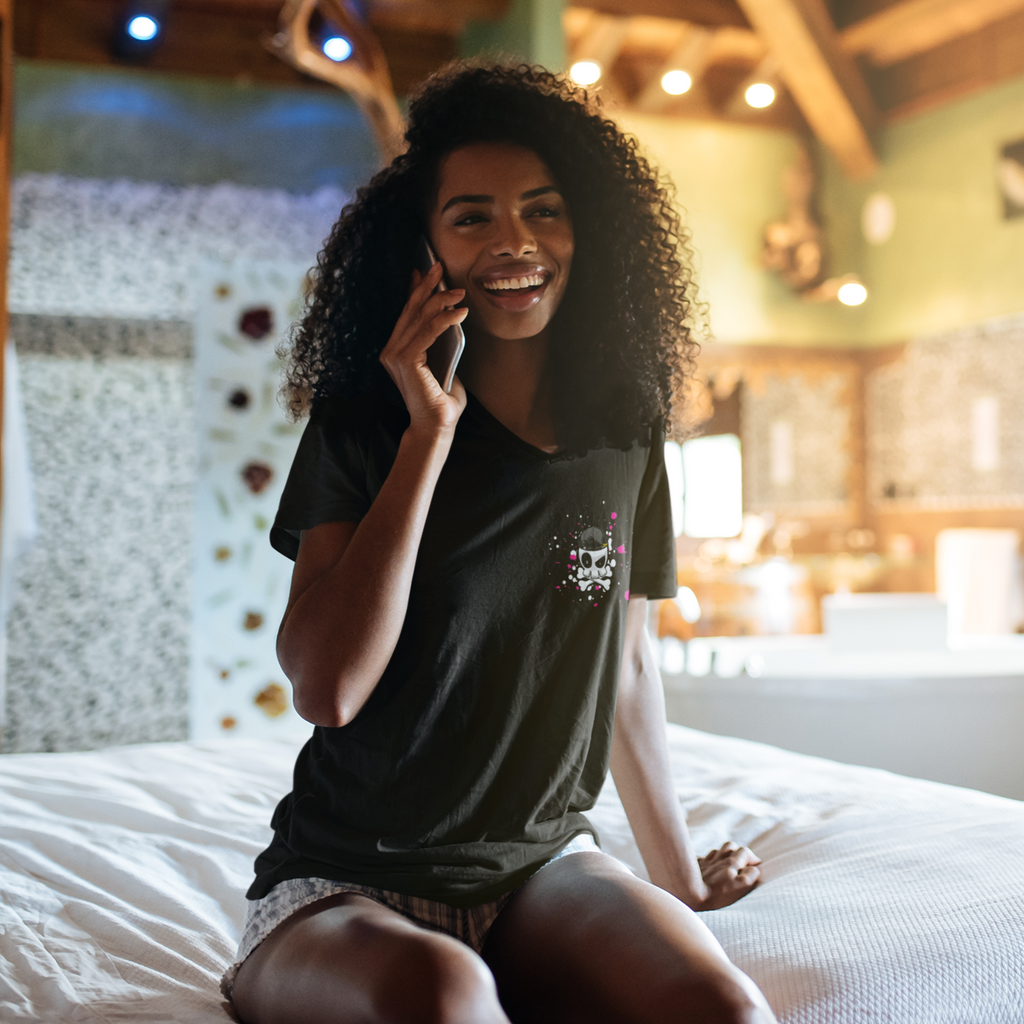 A woman sitting on a bed, talking on her cell phone wearing a black  vneck tshirt with the skull splat design from SlamDannigan